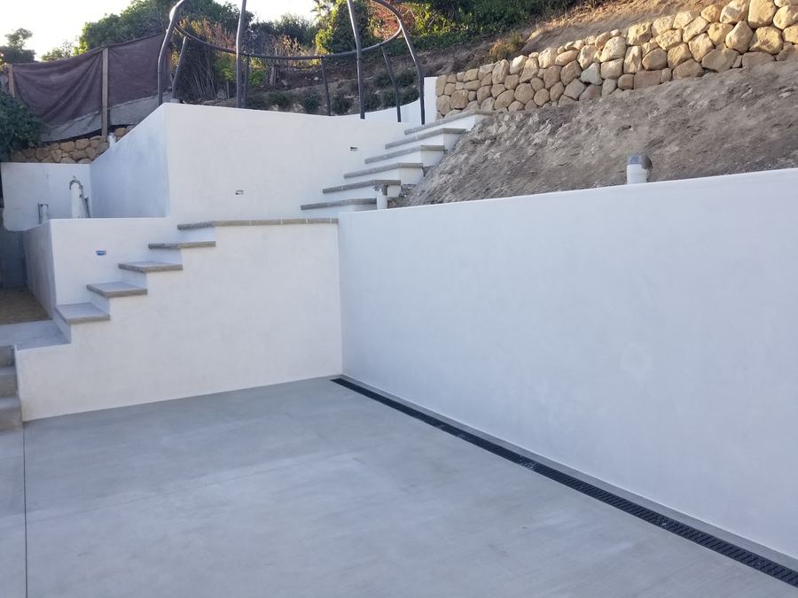 A white stucco garden wall with a concrete pad below it and open stair treads set into the slope.