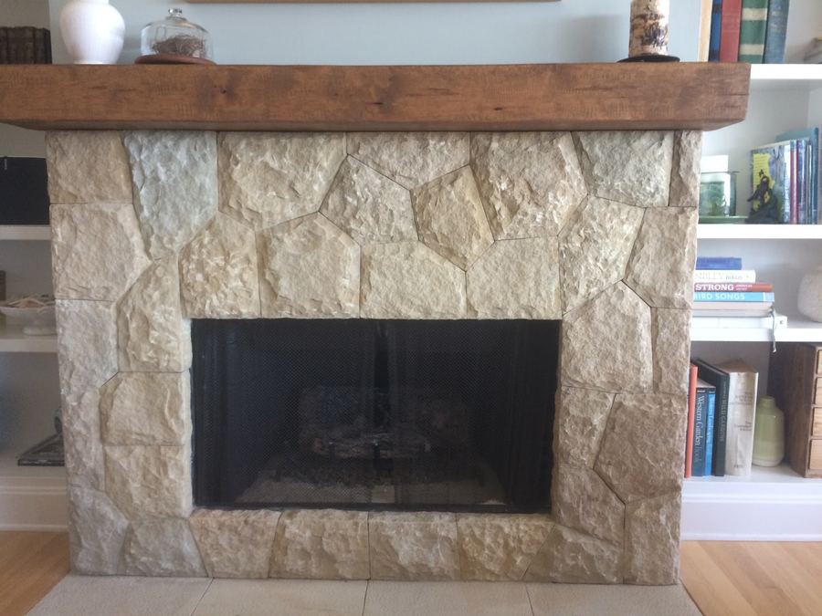 An interior fireplace faced in pale cut stone with a heavy timber mantel and built-in shelving beside it.